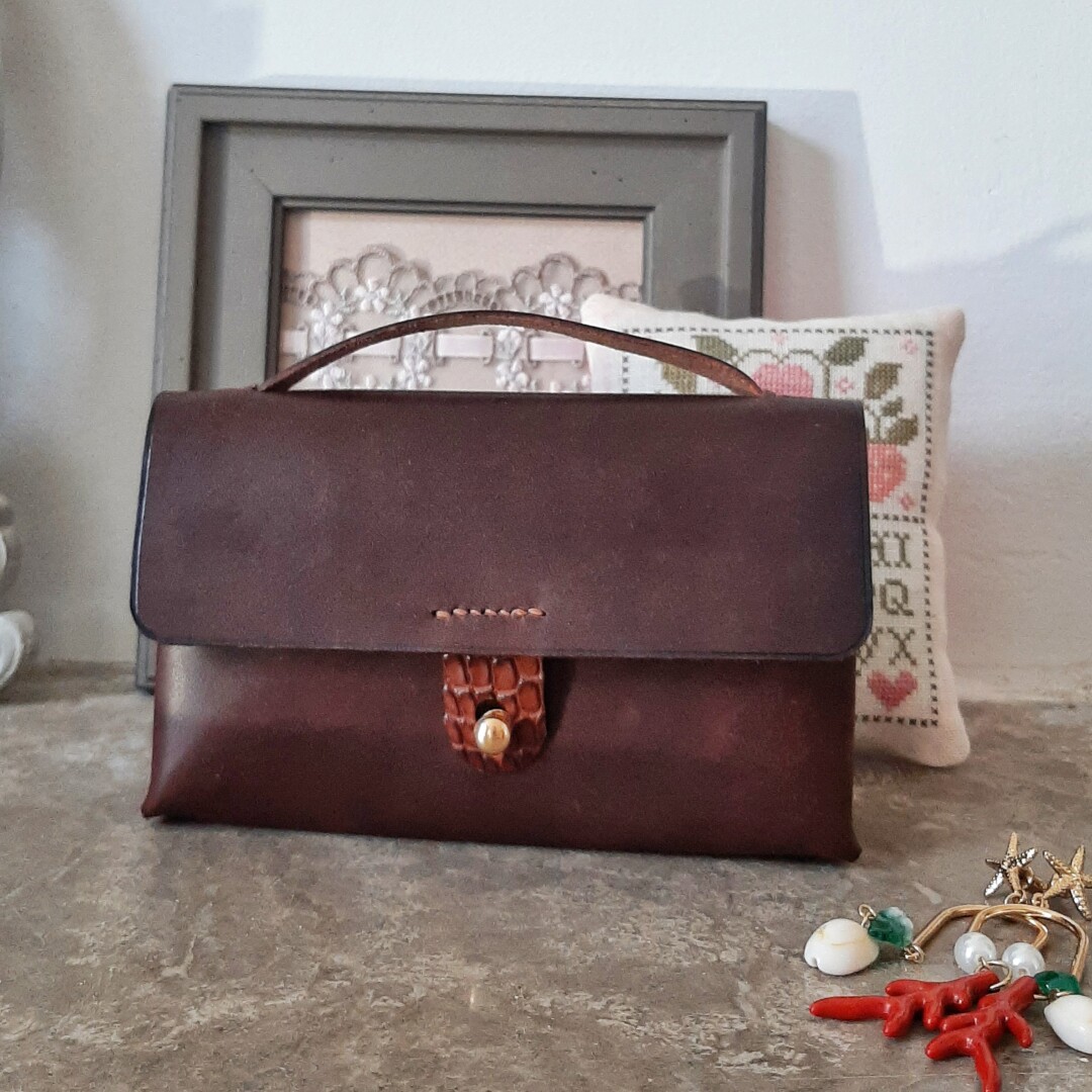 Vue de face de la pochette en cuir lisse tannage végétal de couleur marron chocolat. Fermeture par un bouton de col doré. Languette de fermeture cousue sur le rabat en alligator marron brillant. Poignée en cuir lisse tannage végétal marron, sur le haut pour un transport plus facile