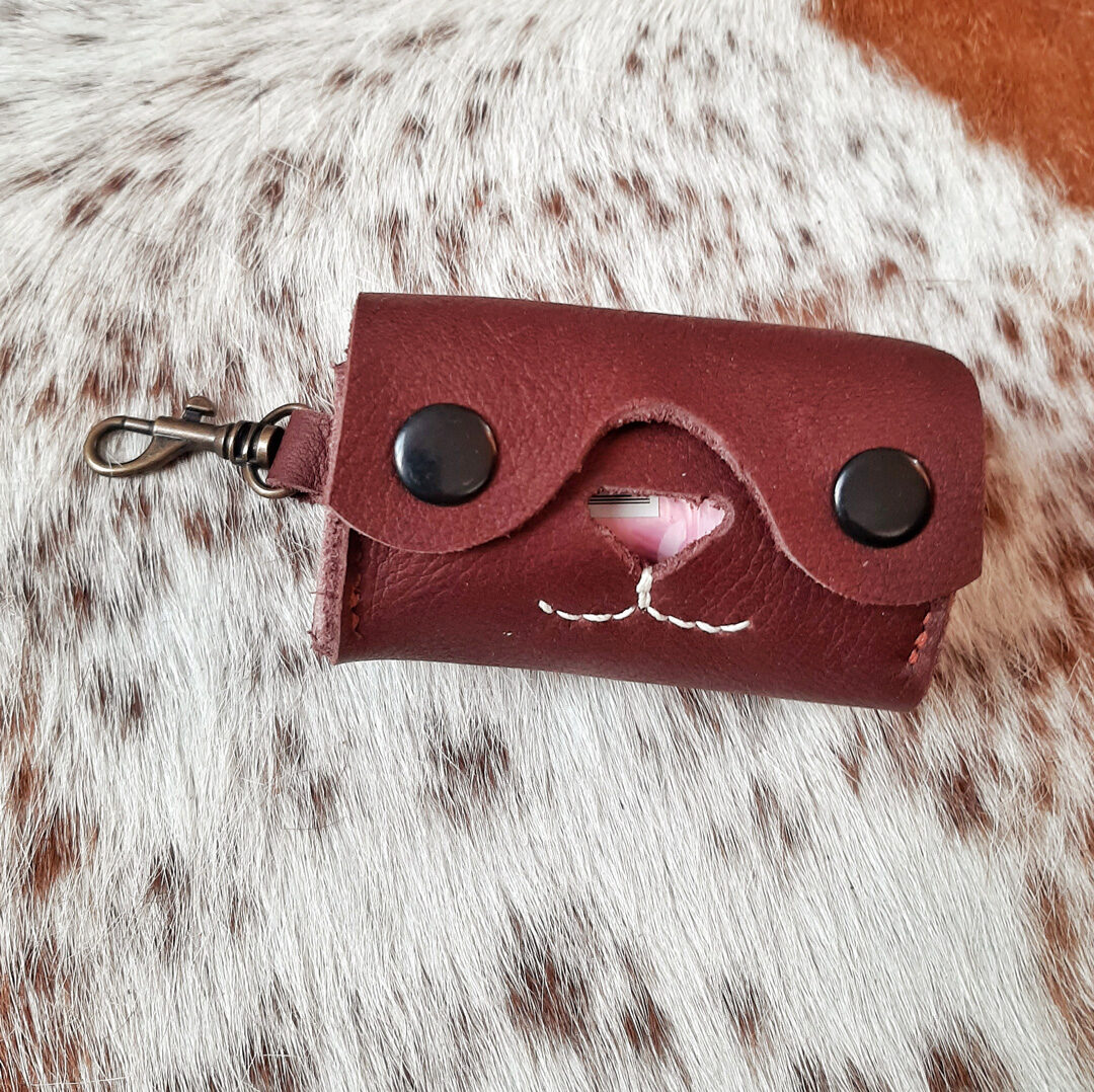 Un porte-sac à crottes élégant en cuir marron chocolat, avec une découpe en forme de cœur révélant un sac en plastique. Le design est épuré, avec deux boutons pression noirs et des coutures apparentes. Un mousqueton en métal suggère une fixation facile à une laisse ou un sac. L'ensemble est présenté sur un fond de peau de vache naturelle, soulignant le côté artisanal du produit.