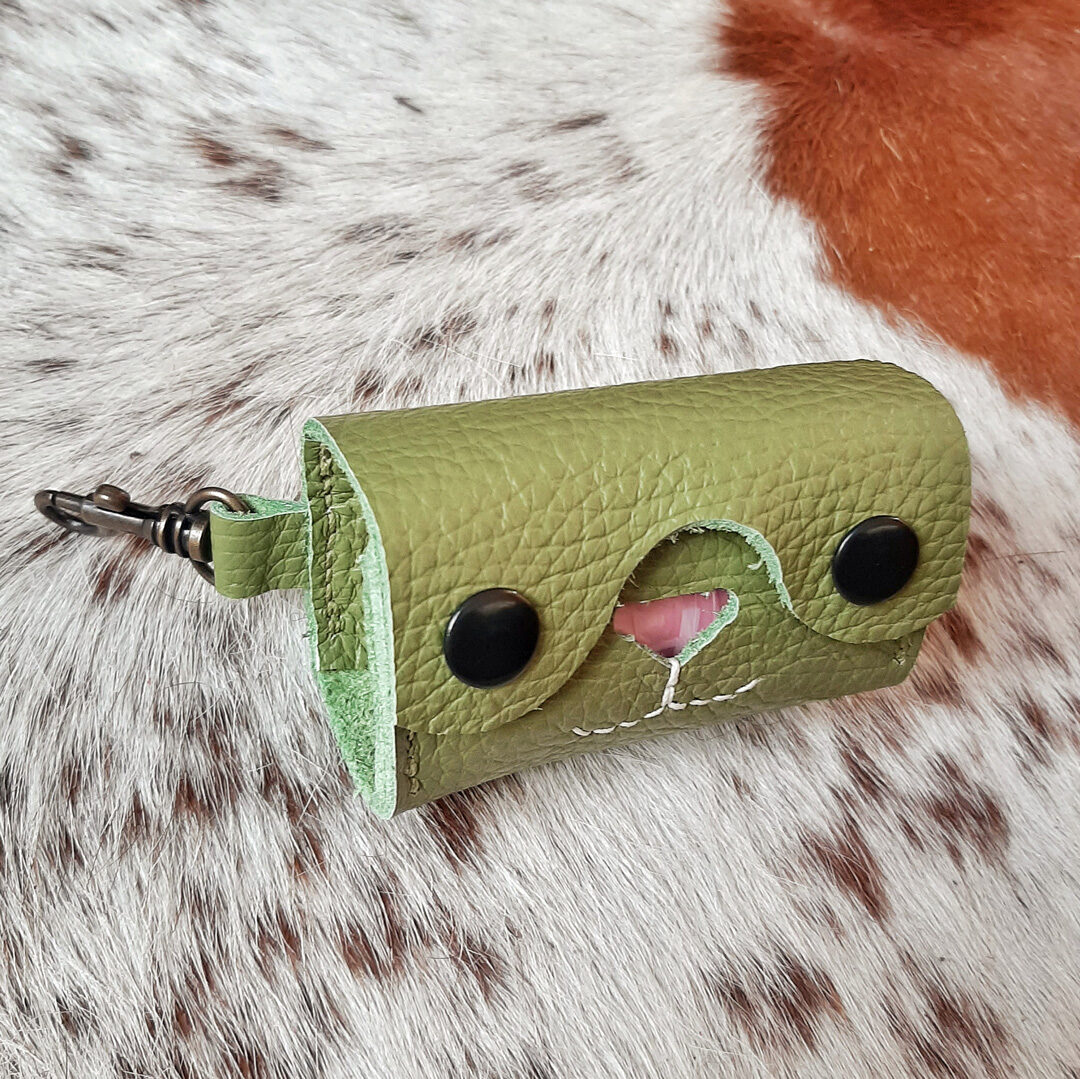 Un porte-sac à crottes élégant en cuir vert amande, avec une découpe en forme de cœur révélant un sac en plastique. Le design est épuré, avec deux boutons pression noirs et des coutures apparentes. Un mousqueton en métal suggère une fixation facile à une laisse ou un sac. L'ensemble est présenté sur un fond de peau de vache naturelle, soulignant le côté artisanal du produit.