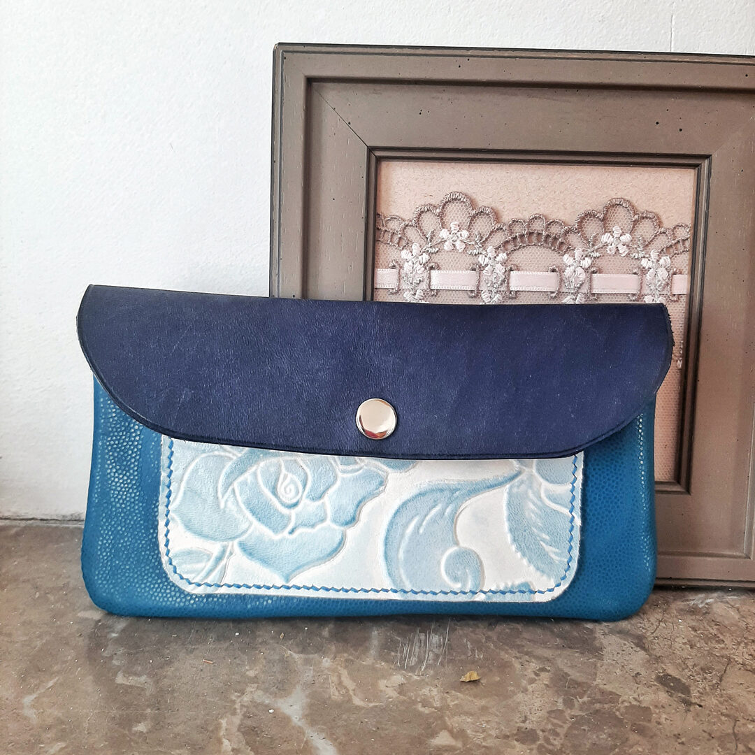 Pochette en cuir tannage végétal bleu, Poche cousu main en cuir tannage végétal, embossé de fleurs bleu turquoise sur fond blanc sur le devant. Le cuir du devant est un cuir fantaisie brillant turquoise embossé imitation grain caviar. Se ferme par un bouton pression argenté.