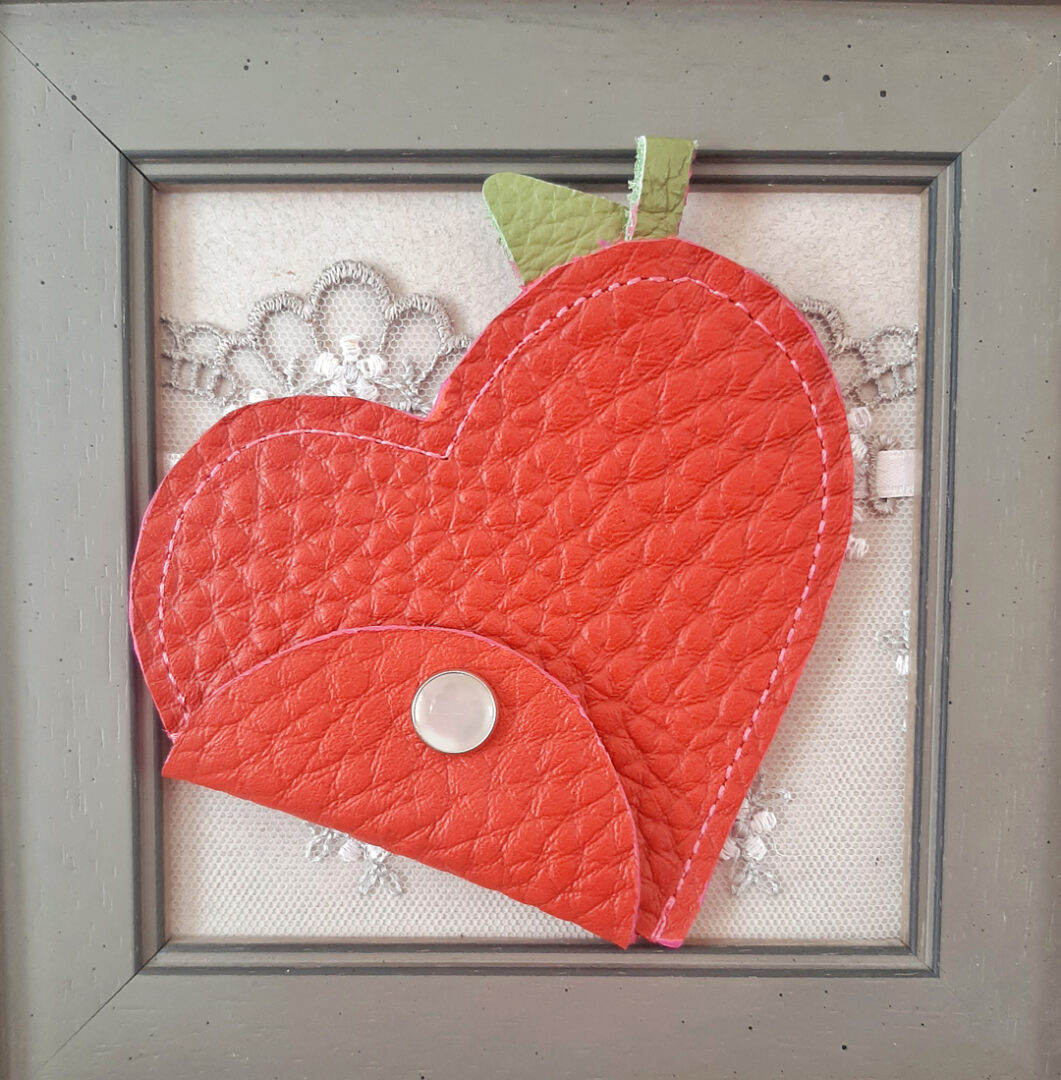 Porte-monnaie en forme de cœur. Rabat se fermant par un bouton pression de couleur nacré. Cuir de luxe rouge grainé dit grain de café. Couture machine de couleur rose, finition main. Tige et feuille en cuir vert, qui représentent celle d'un fruit. intérieur brut
