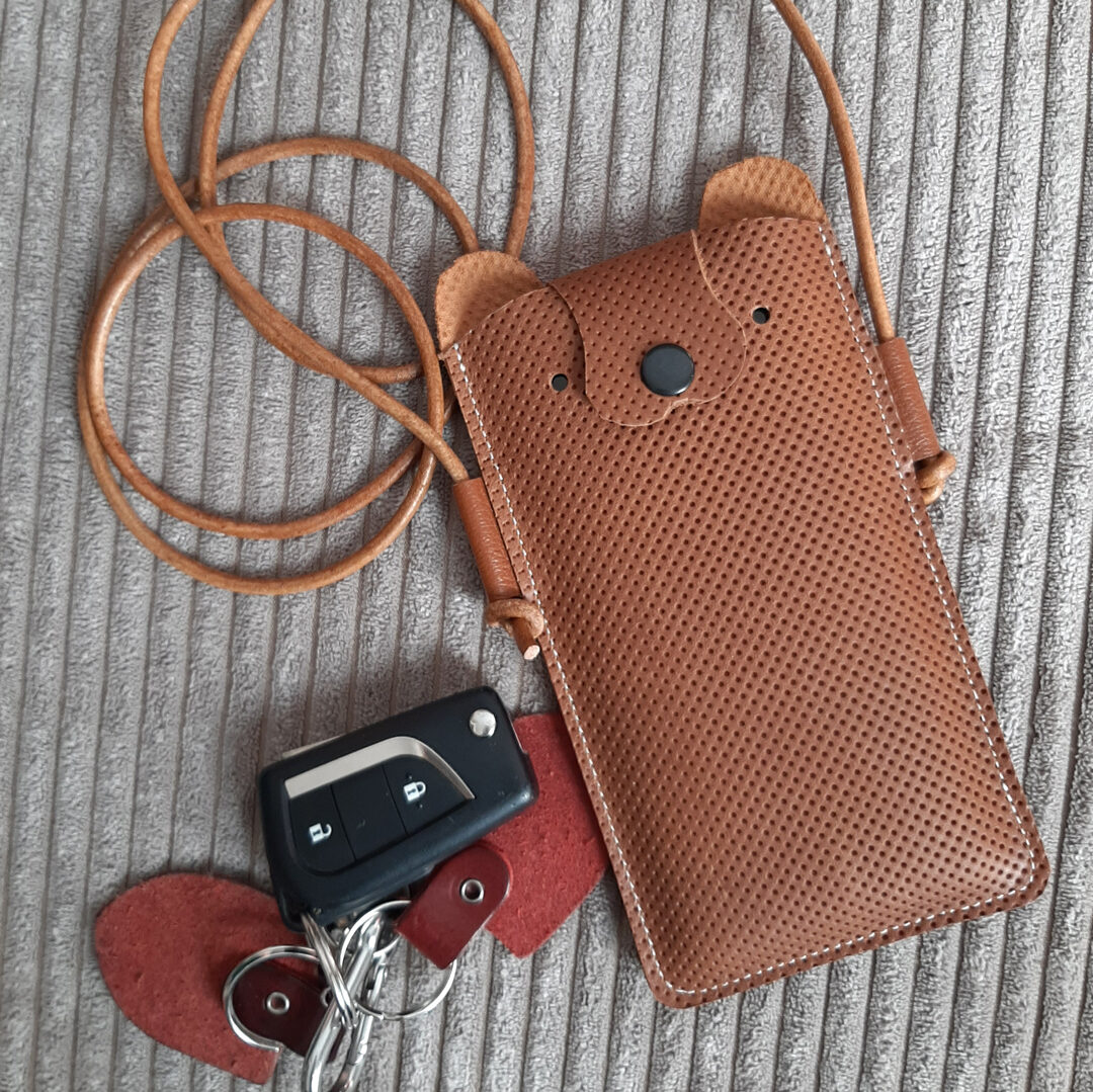 Pochette à portable avec le haut en forme de tête de nounours. Cuir fantaisie perforé sur toute la surface. Joli marron noisette et fermeture par bouton pression noir qui symbolise le nez de l'animal. Cordelette en cuir réglable pour la bandoulière.