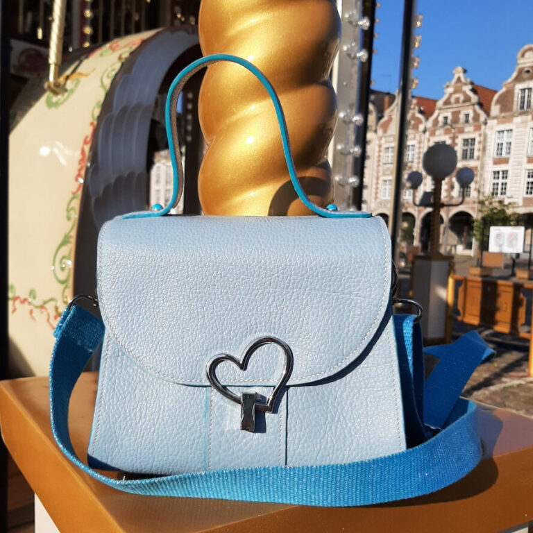 Sac à main en cuir grainé bleu ciel, posé sur un élément doré d’un manège en bois, en extérieur. Le sac est muni d’une bandoulière bleu roi et d’un fermoir en métal argenté en forme de cœur. L’arrière-plan montre une place pavée avec des bâtiments anciens.