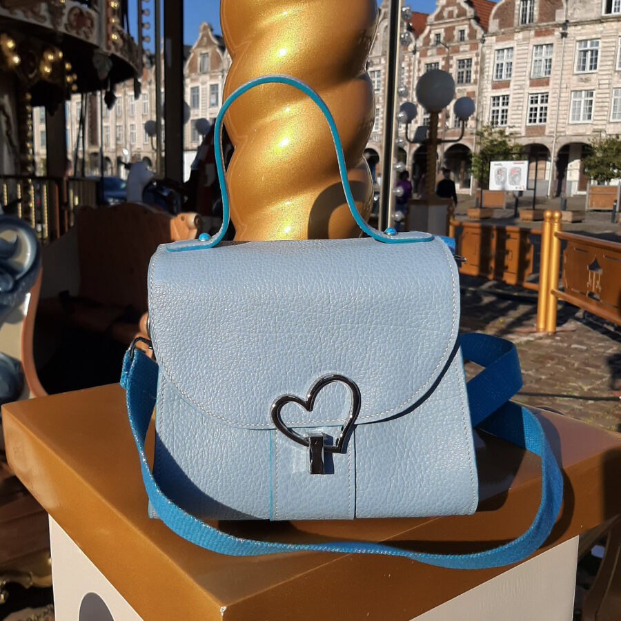 Sac à main en cuir grainé bleu ciel, posé sur un élément doré d’un manège en bois, en extérieur. Le sac est muni d’une bandoulière bleu roi et d’un fermoir en métal argenté en forme de cœur. L’arrière-plan montre une place pavée avec des bâtiments anciens de Arras