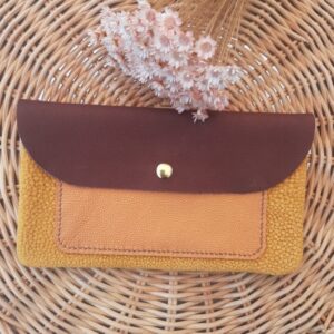 Pochette artisanale en cuir avec rabat bordeaux, poche extérieure en cuir fantaisie irisé grain caviar orange et corps en cuir fantaisie velours grain imitation galuchat camel, fermée par un bouton pression doré, posée sur un fond en rotin