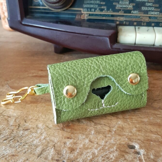 Etui pour rouleau de sachets de sacs à excréments. Cuir pleine fleur vert pistache. Deux boutons pressions dorés, un nez de forme d'un triangle évidé pour laisser passer les sachets en plastique, et une couture cousu main en lin blanc en dessous du nez qui représente les babines d'un chien ou chat. Sur le côté un mousqueton en forme de tete de lapin pour l'accrocher où vous le souhaitez.