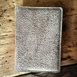 Vue du porte passeport fermé. Cuir fantaisie avec un joli gaufrage de motifs fleuris dans des nuances de marrons glacés et beige, et légèrement irisé. Couture blanc cassé.