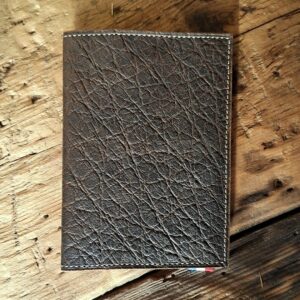 Vue fermé du porte passeport. Cuir dans des nuances de marrons foncés. Gros veinage, couture blanc cassé.