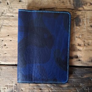 Vue du porte passeport fermé. Cuir lisse, tannage végétal bleu avec impression d'un camouflage dans un camaïeu de bleus. Couture au point sellier, fil de lin bleu turquoise