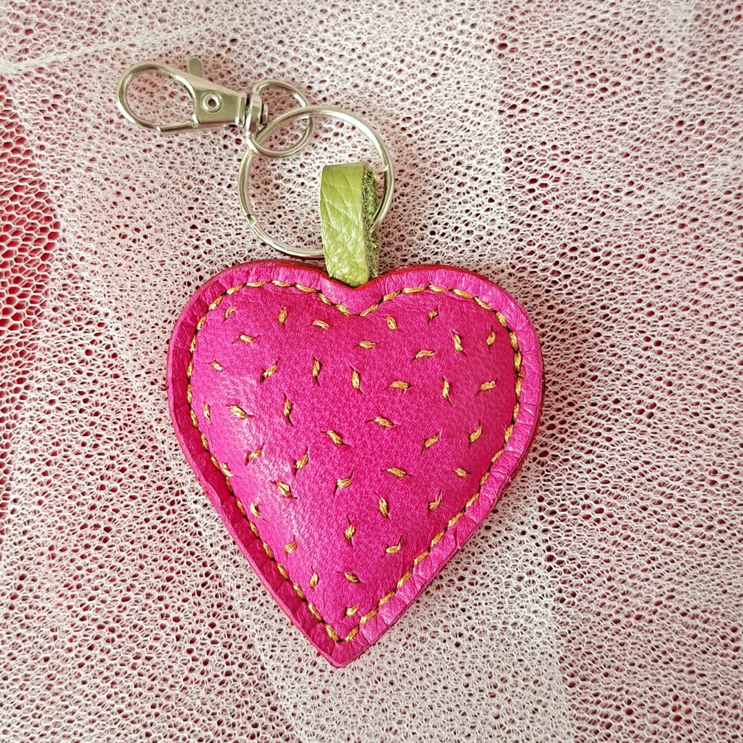 Porte-clés en cuir tannage végétal de couleur rose fuchsia en forme de cœur. Cousu main au point sellier au fil métallisé or. Motif brodé grain de sable à la main avec du fil métallisé or. Mousqueton argenté