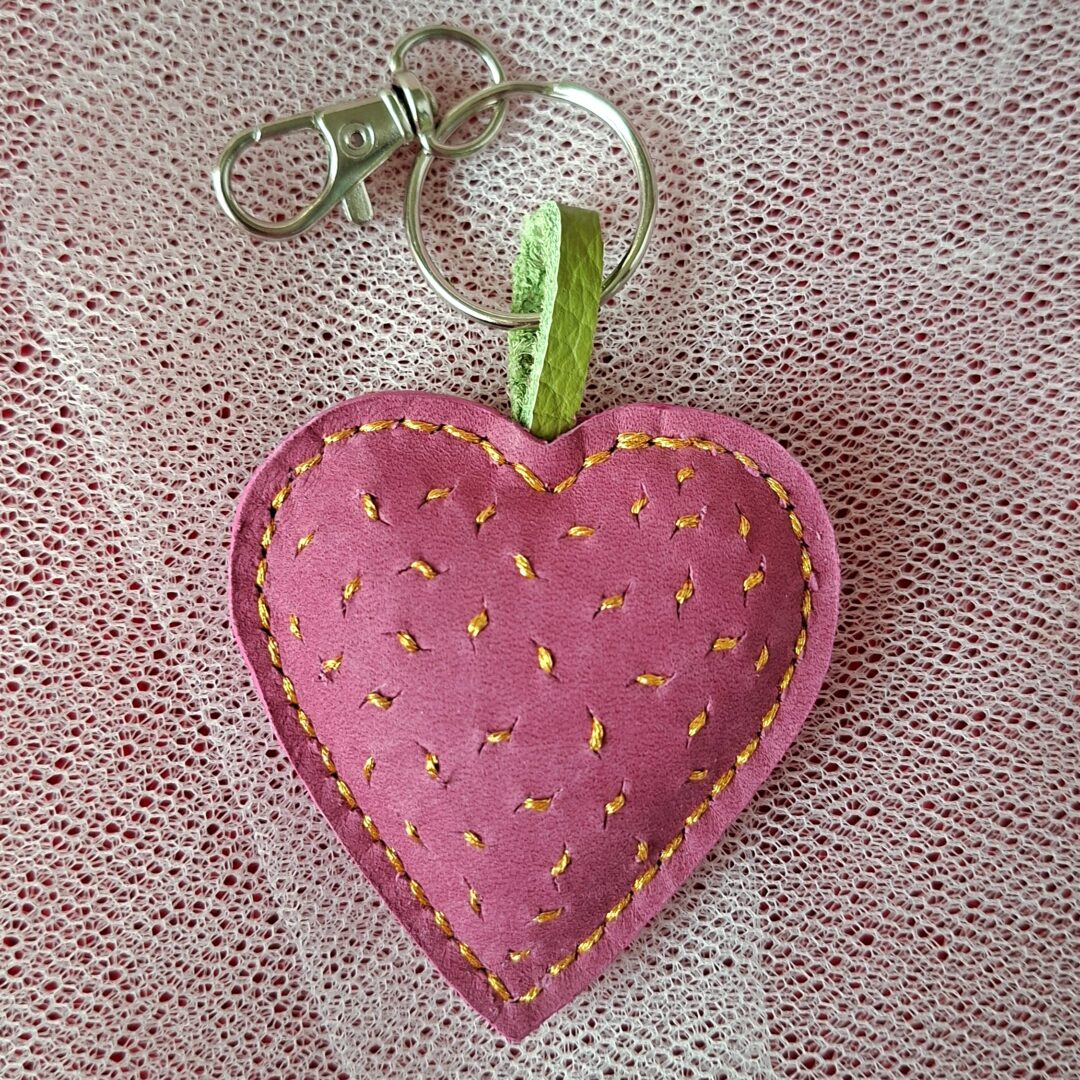 Porte-clés en cuir lisse nubuck rose en forme de cœur. Cousu main au point sellier au fil métallisé or. Motif brodé grain de sable à la main avec du fil métallisé or. Mousqueton argenté