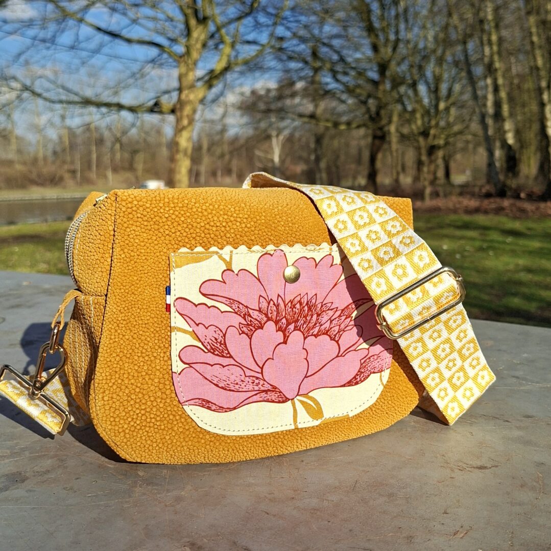 Sac bandoulière style fourre tout, en cuir de chèvre fantaisie de couleur moutarde, touchez velours et imitation d'un effet de motif galuchat. Bandoulière amovible tissé en coton large de 38 mm, aux couleurs assorties au sac. Poche plaquée sur le devant en coton avec un motif d'une grosse fleur rose et feuilles couleur moutarde. Un croquet crème, borde le haut de la poche.